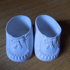 Puede incluir: Un par de zapatos de muñeca en color blanco mate con un detalle de lazo moldeado en la parte delantera y un patrón decorativo de borde festoneado en la puntera. Los zapatos tienen una forma redondeada y un diseño abierto en la parte superior.