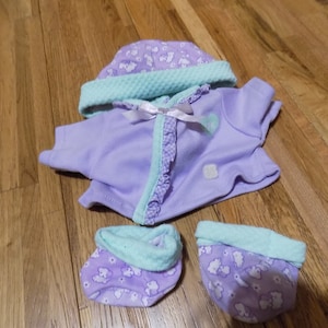 Op de afbeelding: Een paars en groene babypop outfit met een muts, jasje en slofjes. De outfit heeft een bloemenpatroon met witte wolken.