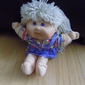 Peut inclure: Une poupée vintage avec un corps en tissu doux et un visage en vinyle. La poupée a les yeux bleu clair, un teint clair et des cheveux gris clair et duveteux. Elle porte une robe bleue à pois blancs et un haut à motifs colorés.