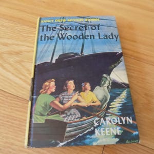May include: Vintage hardcover book titled "The Secret of the Wooden Lady" by Carolyn Keene. The cover features an illustration of three women in a rowboat, a large ship, and a statue. The book has a yellow spine and is set against a wooden background.