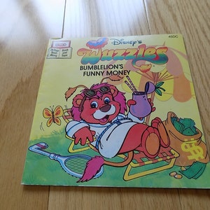 Op de afbeelding: Een vintage Disney's Wuzzles plaat met een cartoon leeuw die een zonnebril en een wit shirt draagt, ontspannen op een strandstoel met een drankje en een tas vol geld. De plaat is getiteld "Bumblelion's Funny Money" en maakt deel uit van de Disney's Wuzzles serie.
