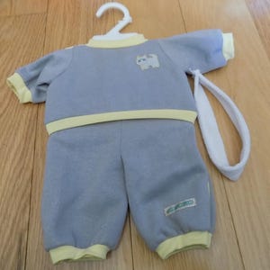 Op de afbeelding: Een grijze en gele babypop outfit met een wit hoofdband. De outfit heeft een kattenontwerp op de voorkant en de tekst "Baby Clothes" op het been.