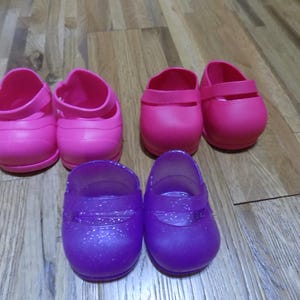 Puede incluir: Seis pares de zapatos infantiles en tonos rosa y morado. Dos pares de zapatos rosas tienen un estilo Mary Jane con una correa. Los otros dos pares son morados con purpurina.