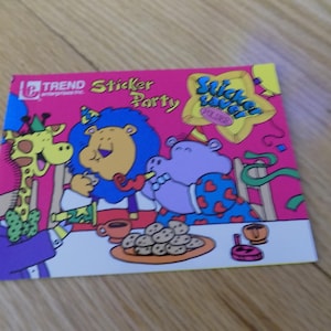 May include: A colorful sticker book titled "Sticker Party" featuring cartoon animals at a party. The cover has a pink background with a giraffe, lion, and hippo. The sticker book is on a wooden surface.
