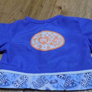 Könnte beinhalten: Blaue und weiße Teddybärjacke mit einem weißen und orangen Patch, auf dem hinten "I love CPK" steht. Die Jacke hat einen weißen Saum und ein blaues und weißes Muster an den Ärmeln und am unteren Rand.
