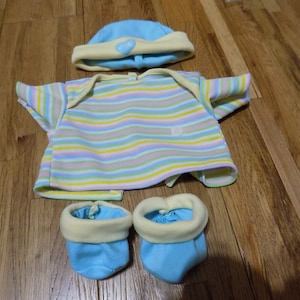 Könnte beinhalten: Ein Baby-Puppen-Outfit mit einem blau-gelb gestreiften Shirt, einer blauen Mütze mit gelbem Besatz und einem Herz mit dem Wort "CIK" darauf und blauen Stiefeln mit gelbem Besatz.