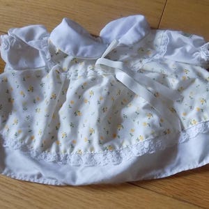 May include: A white doll dress featuring a small yellow floral print, short puffed sleeves, a rounded collar, and a white ribbon tie at the neckline. The garment includes a white lace trim along the hem and a gathered waistline design.