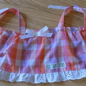 May include: A sleeveless top featuring a pink, orange, and white gingham plaid pattern. The garment has a ruffled white eyelet lace trim along the bottom hem, two shoulder straps with tie closures, and a small white bow at the centre neckline.