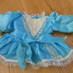 Op de afbeelding: Een lichtblauwe poppenjurk met witte kanten rand en een bijpassende strik. De jurk heeft pofmouwen, een geplooide witte slab en drie kleine witte knopen. De rok heeft een geplooid ontwerp en de jurk is gemaakt van een glanzende stof.