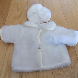 May include: A white plush baby coat and matching bonnet set. The coat has a button-up front, a collar, and fluffy trim along the hem and cuffs. The bonnet is round and fluffy, designed to match the coat.