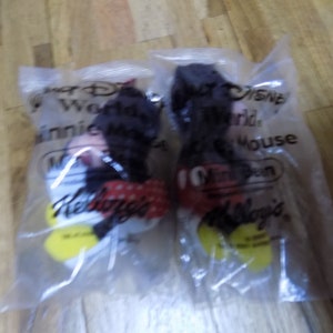 May include: Two plastic bags containing Mickey Mouse plush toys. The bags are labeled "Walt Disney World" and "Mickey Mouse". The bags are also labeled "Kellogg's".