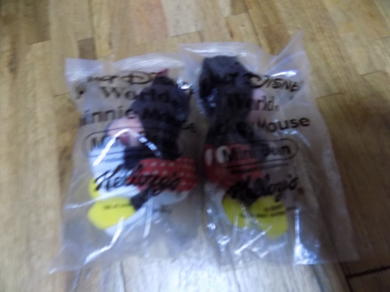 May include: Two plastic bags containing Mickey Mouse plush toys. The bags are labeled "Walt Disney World" and "Mickey Mouse". The bags are also labeled "Kellogg's".