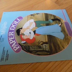 May include: A blue and pink paper doll book featuring Ariel and Eric from Disney's The Little Mermaid. The cover shows Ariel and Eric dancing together. The book includes precut fashions and two dolls. No scissors are needed.