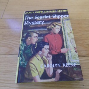 May include: A vintage hardcover book titled "The Scarlet Slipper Mystery" by Carolyn Keene. The cover features a colorful illustration of three people looking at a framed picture. The book is set against a wooden background.