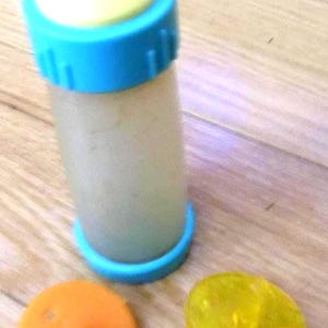 May include: A baby bottle with a blue cap and base, a yellow nipple, and an orange disc. The bottle is filled with a light-colored liquid. The bottle is on a wooden surface.
