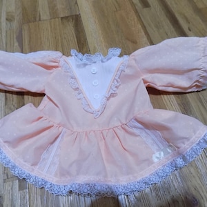 May include: A peach-coloured doll dress with white polka dots, long sleeves, and a ruffled skirt. The dress features white lace trim on the collar, sleeves, and hem, with three white buttons down the front.