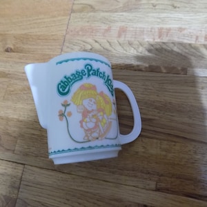 Può includere: Brocca bianca con un design verde e bianco con una bambola Cabbage Patch Kid. La brocca ha un manico e dice "Cabbage Patch Kids" sul davanti.