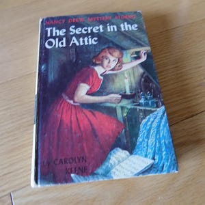 May include: Vintage book cover for "The Secret in the Old Attic" by Carolyn Keene. The cover art depicts a woman in a red dress holding a candle in a dimly lit attic. The book is part of the Nancy Drew Mystery Stories series.