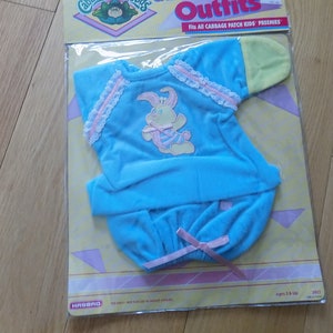 Puede incluir: Un conjunto azul para Cabbage Patch Kids con un diseño de conejo. El conjunto está empaquetado en una bolsa de plástico con un fondo amarillo y rosa. El paquete dice "Outfits" y "Fits all Cabbage Patch Kids Preemies".