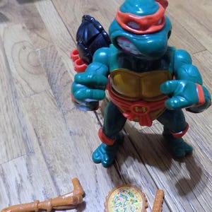 May include: A green Teenage Mutant Ninja Turtles action figure with an orange bandana and a black shell. The figure is holding a pair of sai and has a pizza-shaped weapon on the ground.