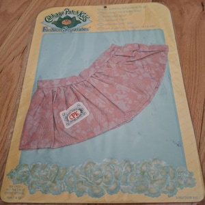 May include: A pink and white floral skirt for a Cabbage Patch Kid doll. The skirt is packaged on a blue background with a Cabbage Patch Kids logo and the words "Fashion Separates".