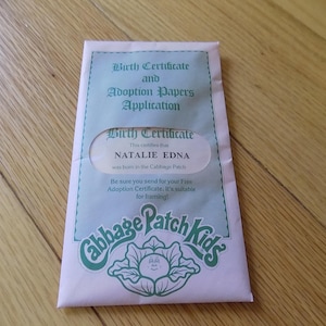 May include: A light pink envelope labelled Cabbage Patch Kids containing a Birth Certificate and Adoption Papers Application. The document inside is visible through a cutout window and displays the name Natalie Edna, stating she was born in the Cabbage Patch.