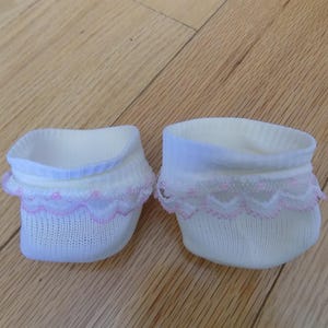 Puede incluir: Dos pares de calcetines tobilleros blancos con un delicado ribete de encaje rosa. Los calcetines están hechos de un material suave y tejido y tienen un ribete azul claro alrededor de la abertura. Están colocados sobre una superficie de madera.