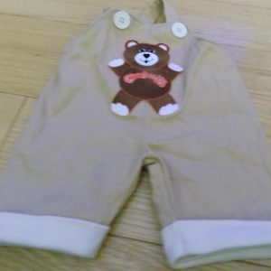 May include: Tan colored baby overalls with a brown teddy bear applique. The teddy bear has a red bow tie and white eyes.