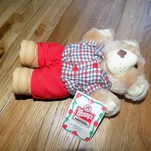Puede incluir: Un oso de peluche marrón que lleva una camisa a cuadros rojos y azules y pantalones rojos. El oso está acostado en un suelo de madera. El oso tiene una etiqueta pegada a su pata que dice "Wendy's Hamburgers".
