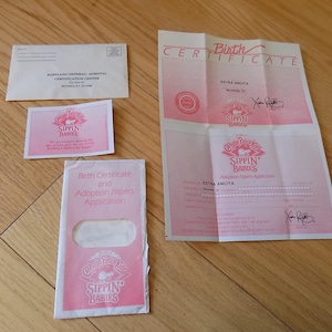 Può includere: Assortimento di documenti cartacei rosa e bianchi, tra cui un certificato di nascita, documenti di adozione e una busta. I documenti presentano il logo e il testo "Sippin' Babies". Gli articoli sono disposti su una superficie di legno.
