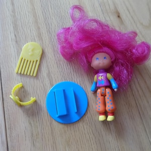 Puede incluir: Una muñeca vintage con cabello rosa brillante, que viste una camisa morada, pantalones naranjas con lunares azules y zapatos amarillos. Los accesorios incluyen un peine amarillo, una pinza para el pelo amarilla y un soporte azul. La muñeca tiene ojos azules y una expresión sonriente.