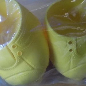 May include: Two bright yellow plastic doll shoes with laces and stitching details. The shoes are packaged in a clear plastic bag.