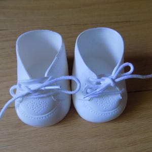 May include: A pair of small, solid white shoes with rounded toes and a smooth finish. Each shoe features a decorative embossed pattern on the toe cap and is secured with thin, white fabric laces tied in a bow at the centre of the opening.