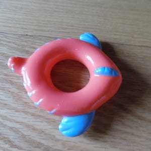 May include: A coral-colored, fish-shaped baby toy with a circular center. The toy has blue fins and accents. The toy is made of a smooth, glossy material and is designed for infants. The toy is lying on a wooden surface.