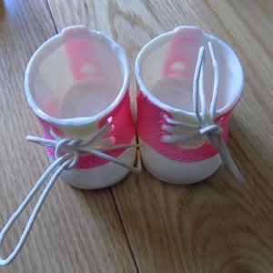 Puede incluir: Un par de zapatos de muñeca de plástico rosa y blanco con cordones blancos. Los zapatos tienen una punta redonda y un pequeño tacón.
