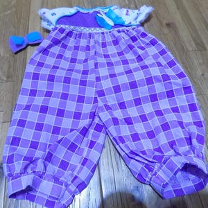 May include: A purple and white checkered baby outfit with a matching bow. The outfit has a white top with a floral pattern and a purple bodice. The pants are a checkered pattern in shades of purple. The bow is a solid purple color.