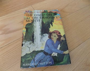 Op de afbeelding: Een vintage boek met de titel "The Discovery at the Dragon's Mouth" van Margaret Sutton. De omslag toont een vrouw in een blauwe jurk bij een waterval en groene rotsformaties. Het boek heeft slijtage.