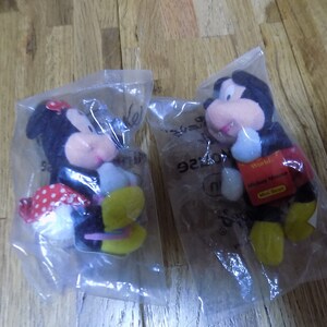 May include: Two plush toys, Mickey Mouse and Minnie Mouse, are packaged in clear plastic bags. Mickey Mouse is wearing red shorts and yellow shoes. Minnie Mouse is wearing a red and white polka dot dress and yellow shoes. The packaging includes the text "World of Disney" and "Mickey Mouse Mini Bean".