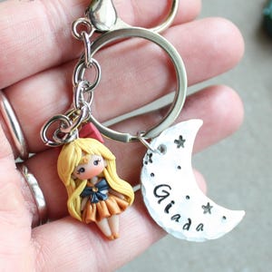 Op de afbeelding: Een handgemaakte sleutelhanger met een kleine pop met blond haar, gekleed in een oranje en blauwe jurk. De sleutelhanger bevat een zilverkleurige sluiting, een ring en een maanvormige bedel met de naam "Giada" en sterren.