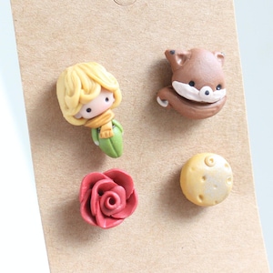 Peut inclure: Un ensemble de cinq petites boucles d'oreilles en argile représentant un renard, une rose, un petit prince et une étoile. Les boucles d'oreilles sont présentées sur une carte brune avec le texte "fait main avec amour".