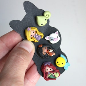 May include: A set of six colorful polymer clay charms featuring Disney princesses and characters. The charms include a green chameleon, a white pig, a yellow fish, a brown-haired princess with a red dress, a black-haired princess with a brown dress, and a blonde princess with a purple dress.