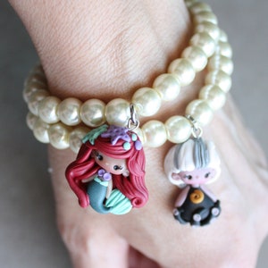 May include: A white pearl bracelet with two charms. One charm is a red-haired mermaid with a green tail and the other is a black and white doll with a black dress.
