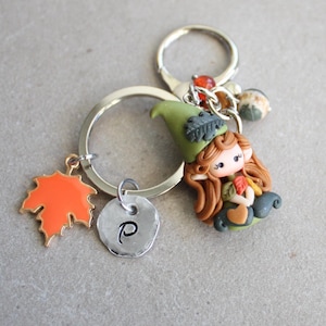 Op de afbeelding: Een zilveren sleutelhanger met herfstthema. Het heeft een kleine kabouterfiguur met een groene hoed, die bladeren vasthoudt, een oranje esdoornbladbedel en een schijf met de letter "P".