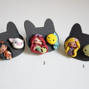 May include: Three sets of handmade polymer clay earrings featuring Disney characters. The first set features a brown-haired girl with a pig, the second set features a red-haired mermaid with a yellow fish, and the third set features a blonde girl with a green chameleon.