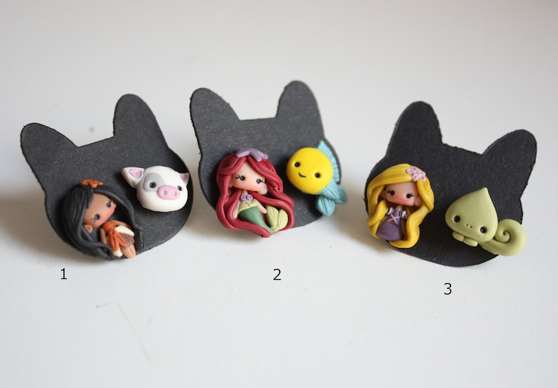 May include: Three sets of handmade polymer clay earrings featuring Disney characters. The first set features a brown-haired girl with a pig, the second set features a red-haired mermaid with a yellow fish, and the third set features a blonde girl with a green chameleon.