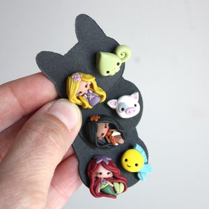May include: A set of six colorful polymer clay charms featuring characters from popular Disney movies. The charms include a green chameleon, a pink pig, a brown-haired girl with a blue dress, a brown-skinned girl with a red dress, a yellow fish with a blue fin, and a red-haired girl with a green tail.
