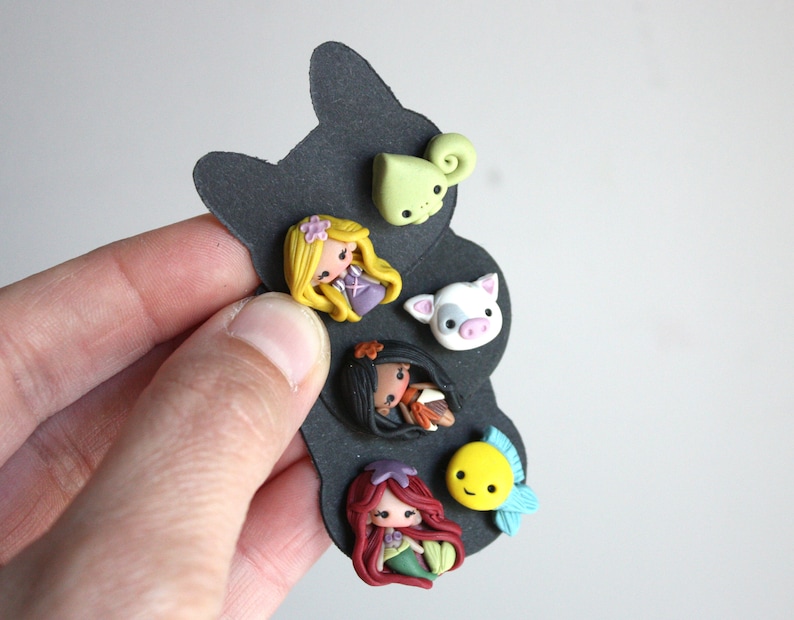 May include: A set of six colorful polymer clay charms featuring characters from popular Disney movies. The charms include a green chameleon, a pink pig, a brown-haired girl with a blue dress, a brown-skinned girl with a red dress, a yellow fish with a blue fin, and a red-haired girl with a green tail.