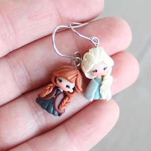 May include: A pair of handmade earrings featuring miniature doll figures. One doll has auburn hair in braids, wearing a dark dress and a burgundy jacket. The other doll has blonde hair in a braid, wearing a light blue dress and jacket.