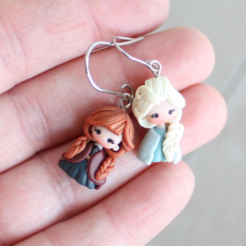 May include: A pair of handmade earrings featuring miniature doll figures. One doll has auburn hair in braids, wearing a dark dress and a burgundy jacket. The other doll has blonde hair in a braid, wearing a light blue dress and jacket.