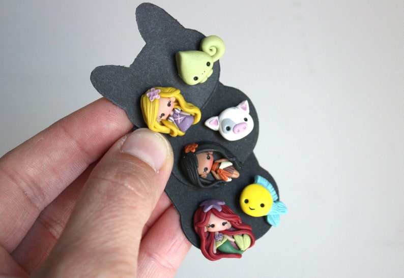 May include: A set of six colorful polymer clay charms featuring Disney princesses and characters. The charms include a green chameleon, a white pig, a yellow fish, a brown-haired princess with a red dress, a black-haired princess with a brown dress, and a blonde princess with a purple dress.